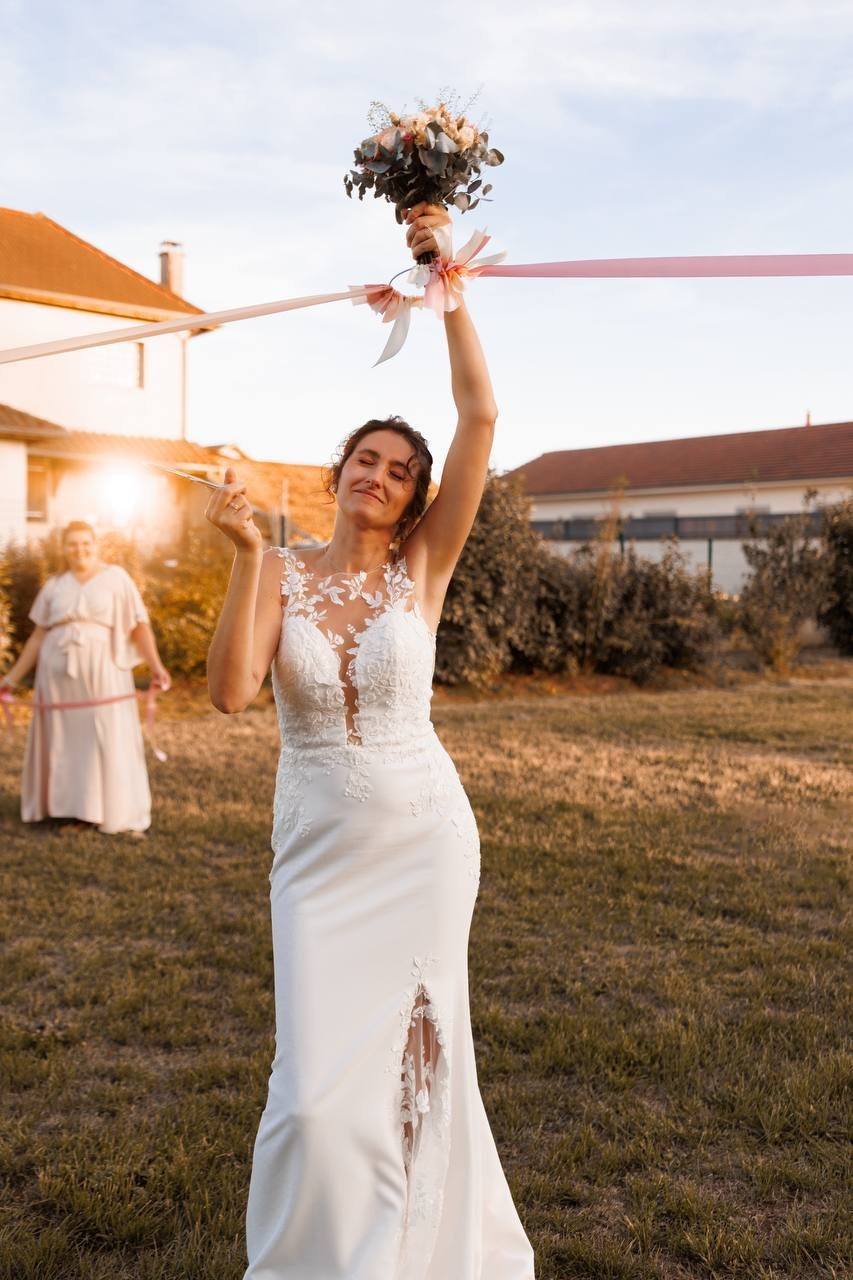 La mariée lève le bouquet à contre-jour, les rubans tendus